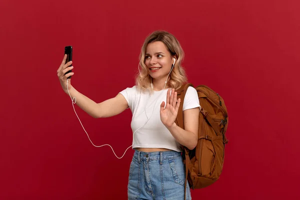 Excited model with curly blond hair dressed in a white t-shirt with orange backpack and white headset video chatting via mobile phone while standing on a red background.