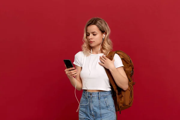 Portrait d'une fille aux cheveux blonds bouclés habillée d'un t-shirt blanc debout sur un fond rouge. Modèle heureux avec sac à dos orange et casque blanc regarde l'écran du téléphone. Concept de voyage — Photo