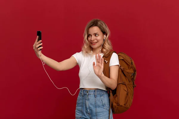 Modelo animado com cabelo louro encaracolado vestido com uma camiseta branca com mochila laranja e fone de ouvido branco conversando por telefone celular enquanto estava em um fundo vermelho . — Fotografia de Stock