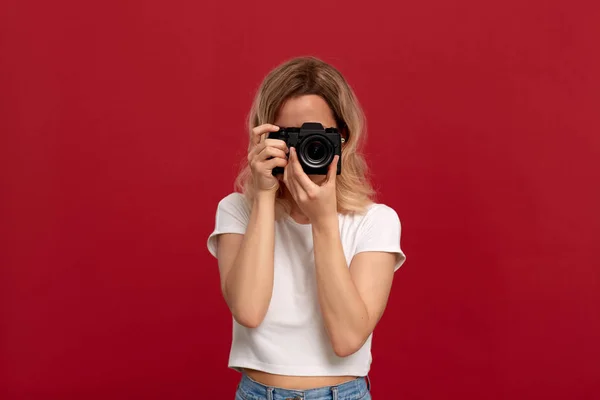 Kırmızı bir arka planda beyaz bir t-shirt ayakta giyinmiş kıvırcık sarı saçlı bir kızın portresi. Model Retro Film kamera ile fotoğraf çeker. 