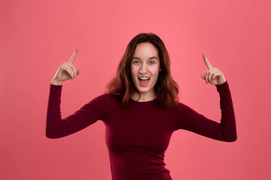 Heyecanlı genç bayan ayakta koyu pembe arka plan üzerinde parmaklarla işaret üzerinde izole. Reklam için yer