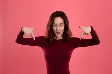 Heyecanlı genç bayan ayakta koyu pembe arka planda parmaklarla aşağı işaret üzerinde izole. Reklam için yer