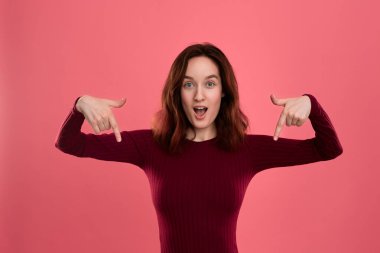 Heyecanlı genç bayan ayakta koyu pembe arka planda parmaklarla aşağı işaret üzerinde izole. Reklam için yer