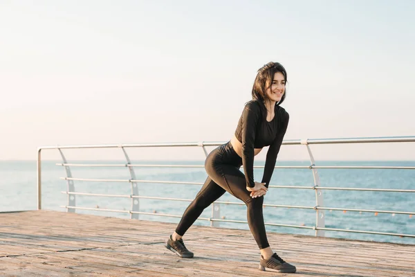 Ung brunett kvinna med vita trådlösa hörlurar i en svart Jumpsuit tar övningar på en brygga vid soluppgången. — Stockfoto