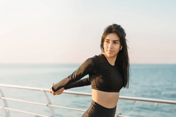 Mujer morena bastante joven en un mono negro se extiende en un muelle al amanecer antes de hacer ejercicio . — Foto de Stock