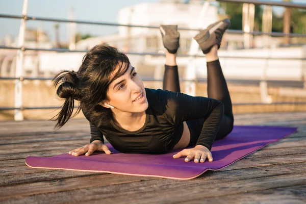 Leende brunett ung kvinna med vita trådlösa hörlurar i en svart Jumpsuit vilar efter plank träning på en matta på piren vid soluppgången. — Stockfoto