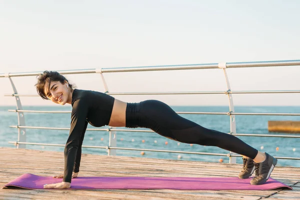 Söt brunett ung kvinna med vita trådlösa hörlurar i en svart Jumpsuit tar plank träning på en matta på piren vid soluppgången. — Stockfoto