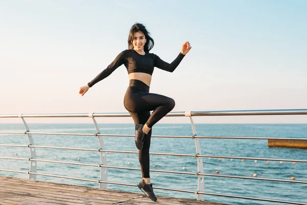 穿着黑色连身衣的快乐年轻黑发女郎在日出时在码头上锻炼。早晨的体育活动,以强健康 — 图库照片