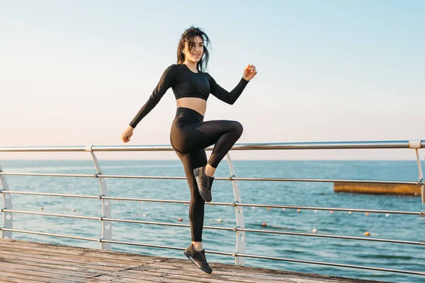 Feliz joven morena en un mono negro toma ejercicios en un muelle al amanecer. Actividad deportiva por la mañana para una salud fuerte — Foto de Stock