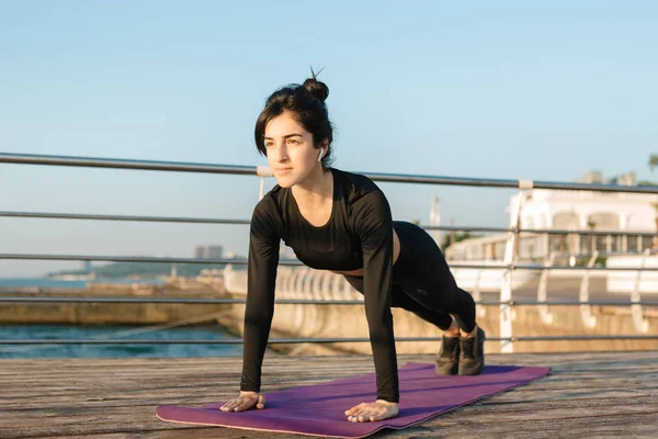 Siyah bir tulum beyaz kablosuz kulaklıklar ile güzel esmer genç kadın güneşin doğuşunda iskelede bir paspas üzerinde tahta egzersiz alır.