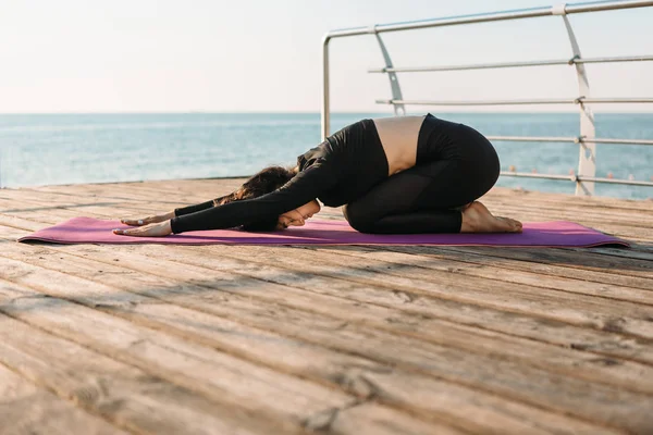 Ung brunett kvinna i en svart Jumpsuit öva yoga stretching övningar på stranden vid soluppgången. Begreppet välbefinnande och hälsosam livsstil. — Stockfoto