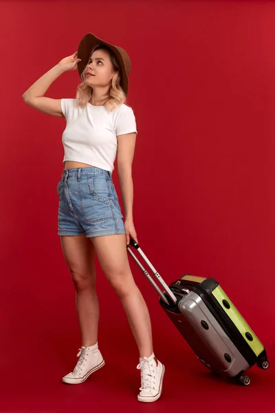 Happy young blond curly woman in a sundown hat holds grey luggage bag while standing infront of a red background — Stock Photo, Image