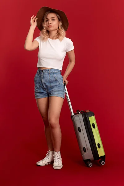 Ung blond lockig kvinna i en Sundown hatt innehar grå bagage påse stående framför en röd bakgrund — Stockfoto