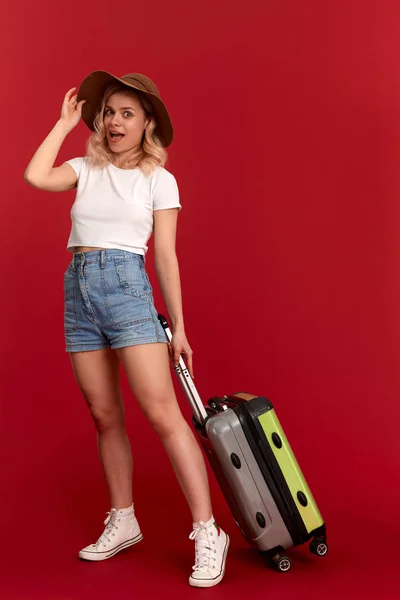 Happy young blond curly woman in a sundown hat holds grey luggage bag while standing infront of a red background