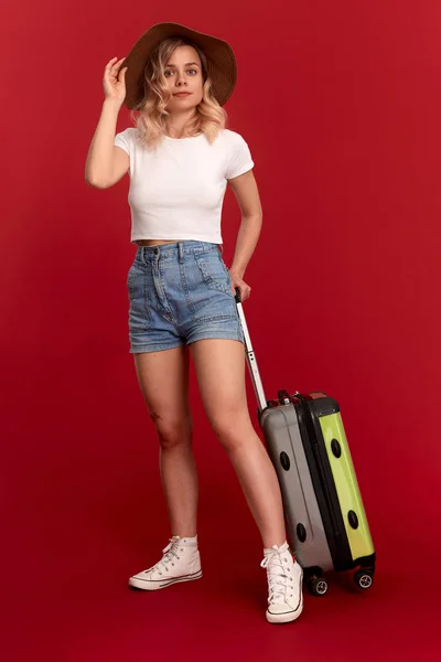 Lycklig ung blond lockig kvinna i en Sundown hatt innehar grå bagage påse stående framför en röd bakgrund — Stockfoto