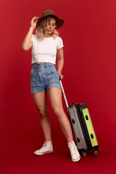 Feliz joven rubia rizada mujer en un sombrero de puesta del sol sostiene bolsa de equipaje gris mientras está de pie delante de un fondo rojo — Foto de Stock