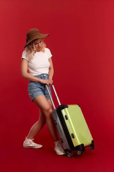 Νεαρή ξανθιά σγουρά γυναίκα με ένα καπέλο στο ηλιοβασίλεμα αισθάνεται κουρασμένοι κρατώντας βαρύ γκρι τσάντα αποσκευών σε περιοδεία αξιοθέατα, ενώ στέκεται μπροστά από ένα κόκκινο φόντο. — Φωτογραφία Αρχείου