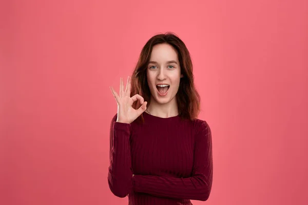 Glückliche junge Frau, die isoliert vor dunkelrosa Hintergrund steht, blickt in die Kamera, während sie eine Handfläche in der Nähe ihres Auges hält und Geste des Ok-Zeichens zeigt. — Stockfoto