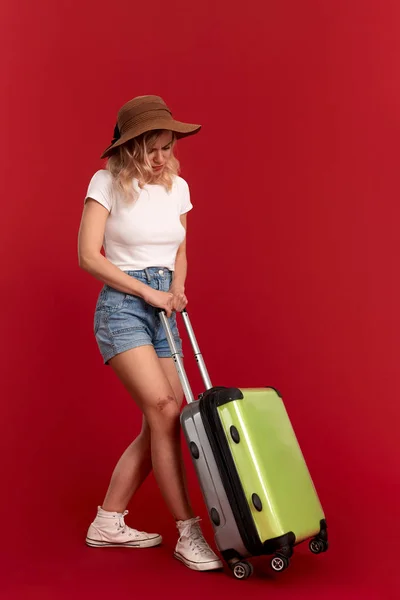 Молодая блондинка в шляпе заката чувствует себя уставшей, держа тяжелую серую сумку для багажа во время экскурсии, стоя перед красным фоном. . — стоковое фото