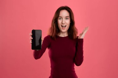 Heyecanlı güzel esmer kız kameraya bakarak koyu pembe bir arka plan üzerinde dururken bir cep telefonu ekran gösteren.
