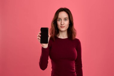 Gülümseyen güzel esmer kız kameraya bakarak koyu pembe bir arka plan üzerinde dururken bir cep telefonu ekran gösteren.