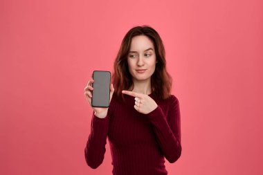 Şüpheci güzel esmer kız koyu pembe arka plan üzerinde dururken bir parmak ile bir cep telefonu ekranında işaret.