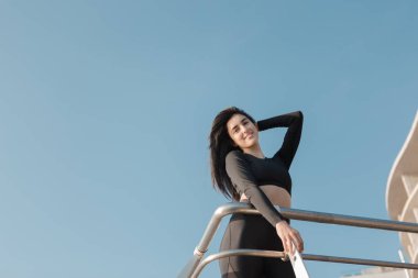 Siyah spor tulum genç güzel esmer kadın kameraya bakarak yoga excercising sonra rahatlatıcı metal heanrails yakınında duruyor