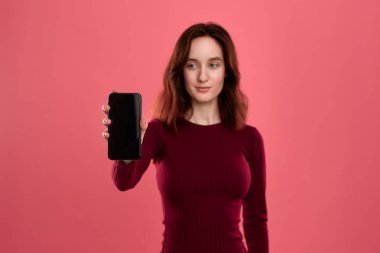 Koyu pembe bir arka plan üzerinde dururken bir cep telefonu ekran gösteren gülümseyen güzel esmer kız.