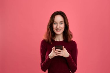 Kamera bakarak koyu pembe bir arka plan üzerinde dururken cep telefonu tutan heyecanlı güzel esmer kız.