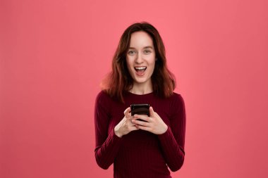 Kamera bakarak koyu pembe bir arka plan üzerinde dururken cep telefonu tutan heyecanlı güzel esmer kız.