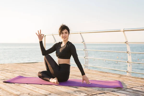 Vacker ung brunett kvinna i en svart Jumpsuit tränar yoga på stranden vid soluppgången. Begreppet välbefinnande och hälsosam livsstil. — Stockfoto