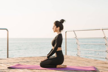 Siyah tulum içinde Gülümseyen esmer genç kadın gün doğumunda iskelede bir paspas üzerinde plank egzersiz sonra dinlenir.