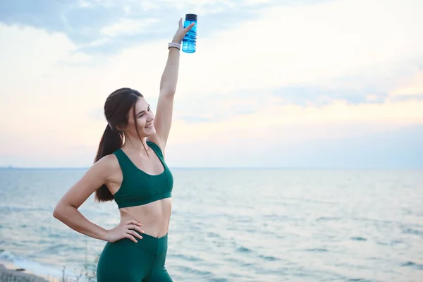 Mujer morena joven con auriculares inalámbricos y relojes inteligentes descansando después del entrenamiento matutino agua potable . — Foto de Stock