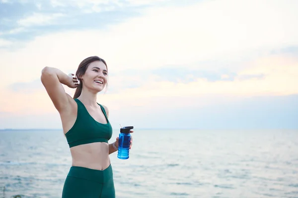 Young brunette woman with wireless earbuds and smart watches resting after morning workout drinking water.