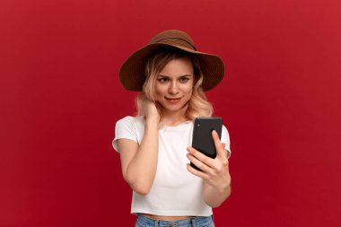 Beyaz bir t-shirt ve günbatımı şapka kıvırcık saçlı sarışın kız kırmızı bir arka plan üzerinde cep telefonu ile selfie yapar.
