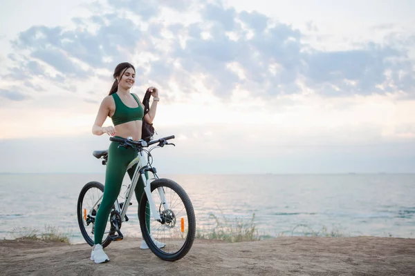 Young brunette woman with a backpack and bicycle rest at the sea shore in the morning after cycling. — Stock Photo, Image