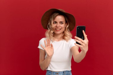 Beyaz bir t-shirt ve günbatımı şapka kıvırcık saçlı sarışın kız kırmızı bir arka plan üzerinde cep telefonu ile selfie yapar.