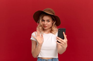 Beyaz bir t-shirt ve günbatımı şapka kıvırcık saçlı sarışın kız kırmızı bir arka plan üzerinde cep telefonu ile selfie yapar.