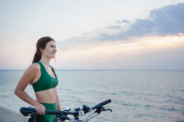 Vacker ung brunett kvinna vilar efter cykeltur vid soluppgången. Modell Lyssna på musik med trådlösa öronsnäckor på morgonen. — Stockfoto