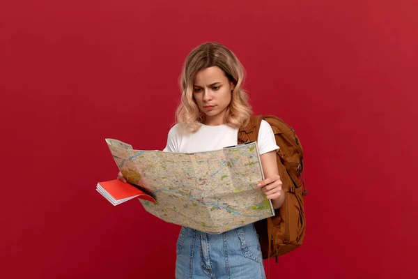 Schockiertes blondes Mädchen mit lockigem Haar in weißem T-Shirt schaut auf die Landkarte und versucht, eine Route mit braunem Rucksack zu finden — Stockfoto