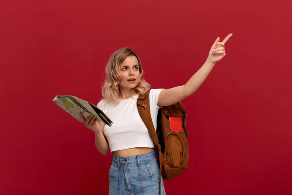 Blond tourist with curly hair in a white t-shirt pointing with a hand at the right during sightseeing tour holding map and brown backpack.