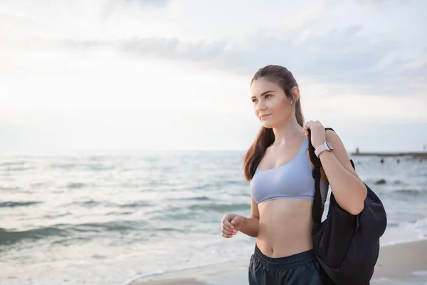 Young brunette woman with wireless earbuds, smart watches and black backpack resting after morning workout at the sea shore at sunrise.