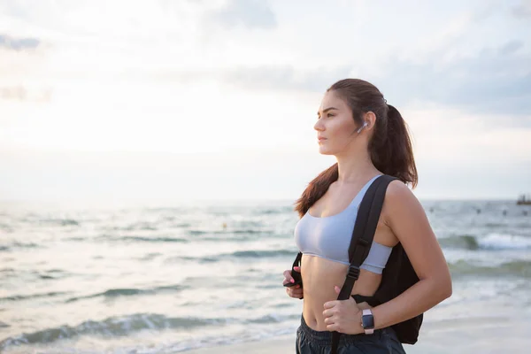 Young brunette woman with wireless earbuds, smart watches and black backpack resting after morning workout at the sea shore at sunrise.