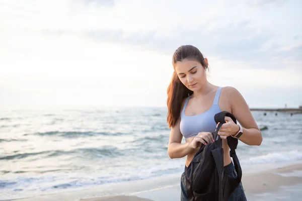 Young brunette woman with wireless earbuds, smart watches and black backpack resting after morning workout at the sea shore at sunrise.