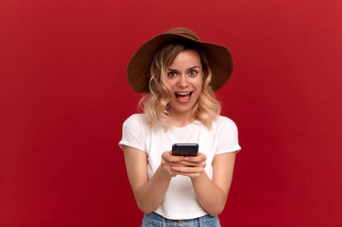 Beyaz bir tişört ve kırmızı arka planda bir güneş ışığı şapka kıvırcık sarı saçlı bir kızın portresi. Mutlu model bir cep telefonu ekranında bakıyor.