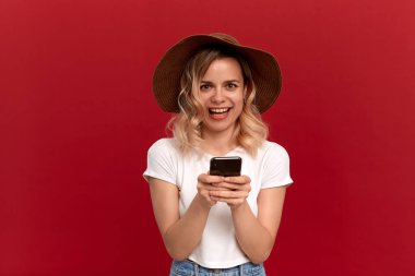 Beyaz bir tişört ve kırmızı arka planda bir güneş ışığı şapka kıvırcık sarı saçlı bir kızın portresi. Mutlu model bir cep telefonu ekranında bakıyor.