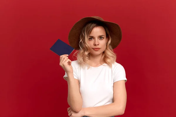 Retrato de una chica con el pelo rubio rizado en una camiseta blanca y un sombrero al atardecer sobre un fondo rojo. Modelo sostiene pasaporte está listo para viajar . —  Fotos de Stock