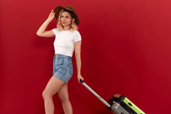 Joven mujer rubia rizada en un sombrero de puesta de sol con bolsa de equipaje gris se emociona en el recorrido turístico mientras está de pie frente a un fondo rojo . —  Fotos de Stock