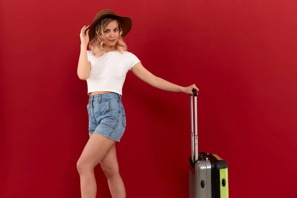 Young blond curly woman in a sundown hat with grey luggage bag is excited at sightseeing tour while standing infront of a red background. — Stock Photo, Image