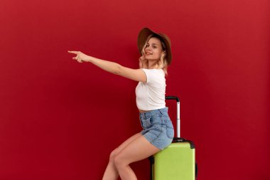 Genç sarışın kıvırcık kadın bir çerçevenin üst kısmında bir parmak ile işaret kırmızı bir arka plan önünde gri bir bagaj çantası üzerinde oturur.